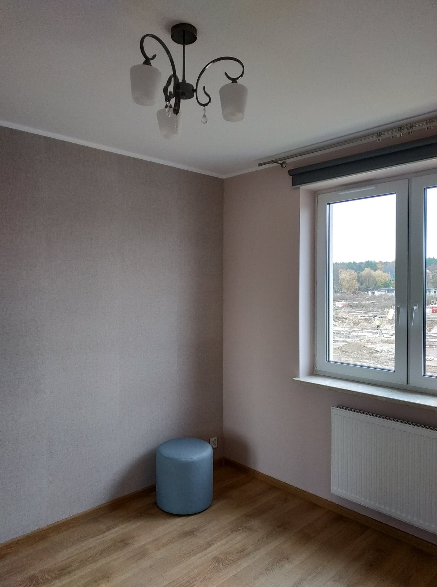 Pokój po remoncie z pomalowanymi ścianami na jasny kolor, drewnopodobną podłogą, grzejnikiem pod oknem i szarą roletą, widok na plac budowy za oknem, oraz lampą sufitową z czarnymi ramionami.