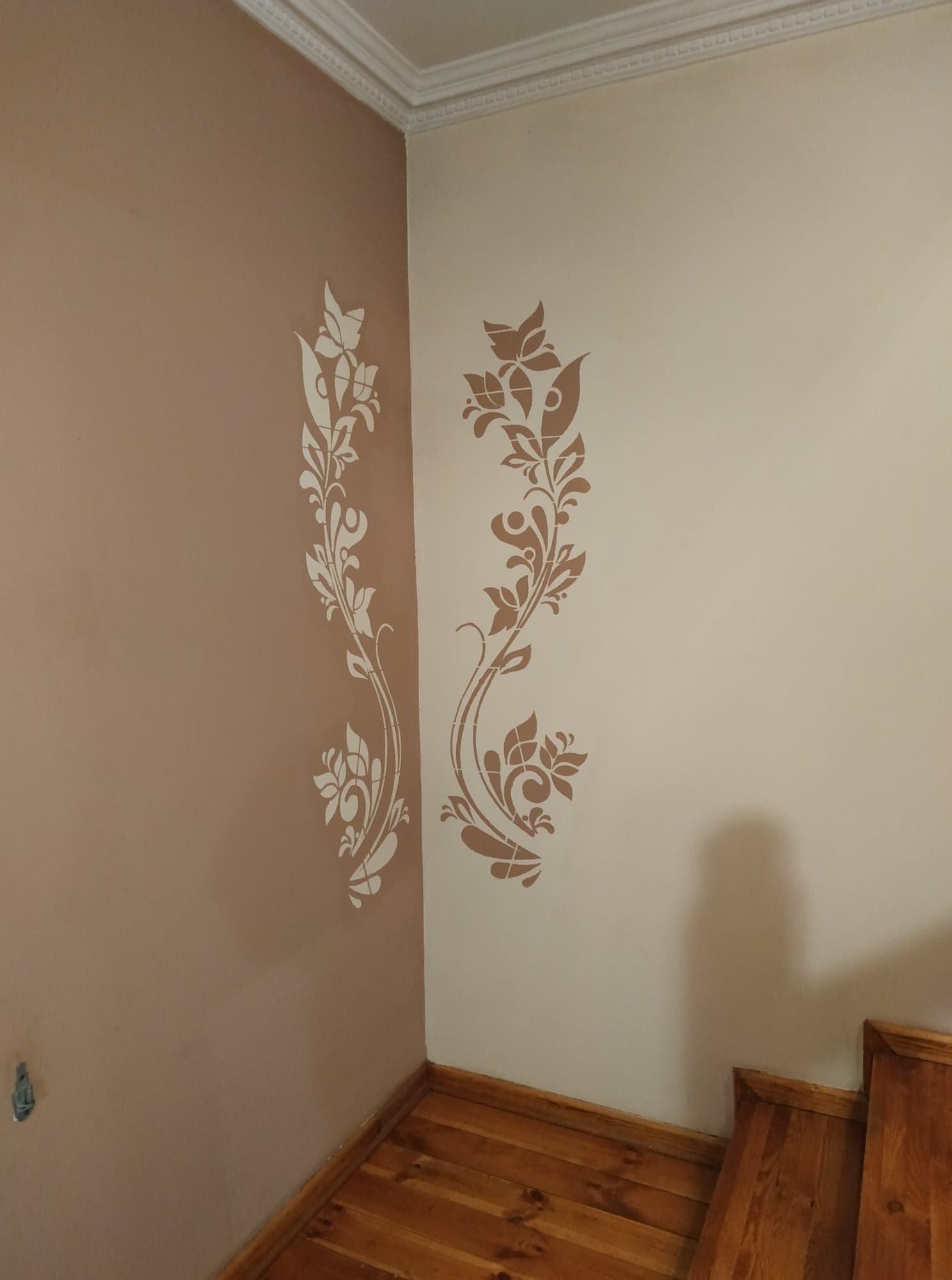 Dekoracyjne malowanie ścian w rogu pomieszczenia: beżowa ściana z białym wzorem roślinnym i kremowa ściana z brązowym motywem roślinnym, widoczne drewniane schody.
