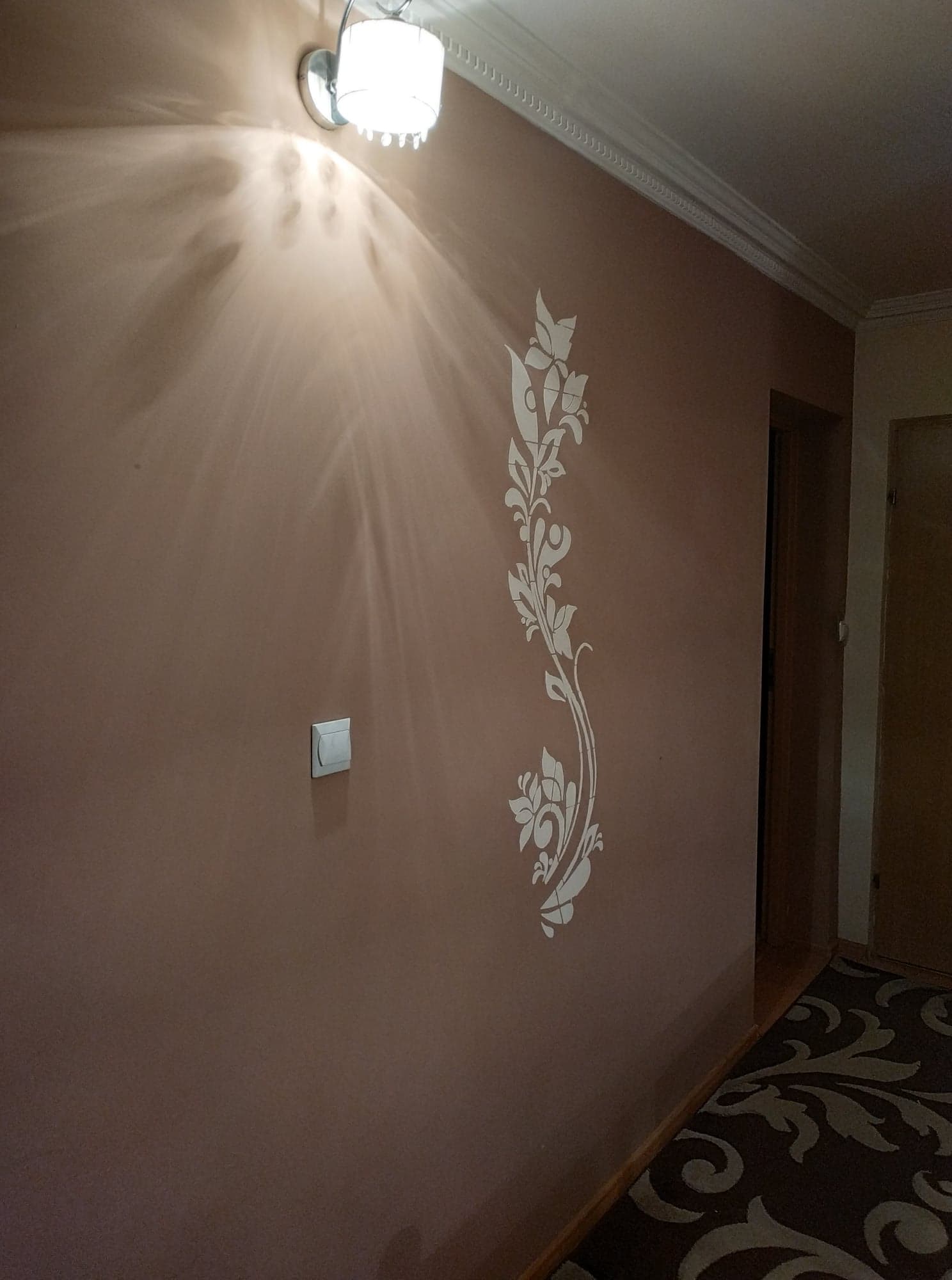 Beżowa ściana z ozdobnym, białym motywem roślinnym, oświetlona lampą sufitową, widoczny włącznik światła i fragment podłogi z wzorzystym dywanem.