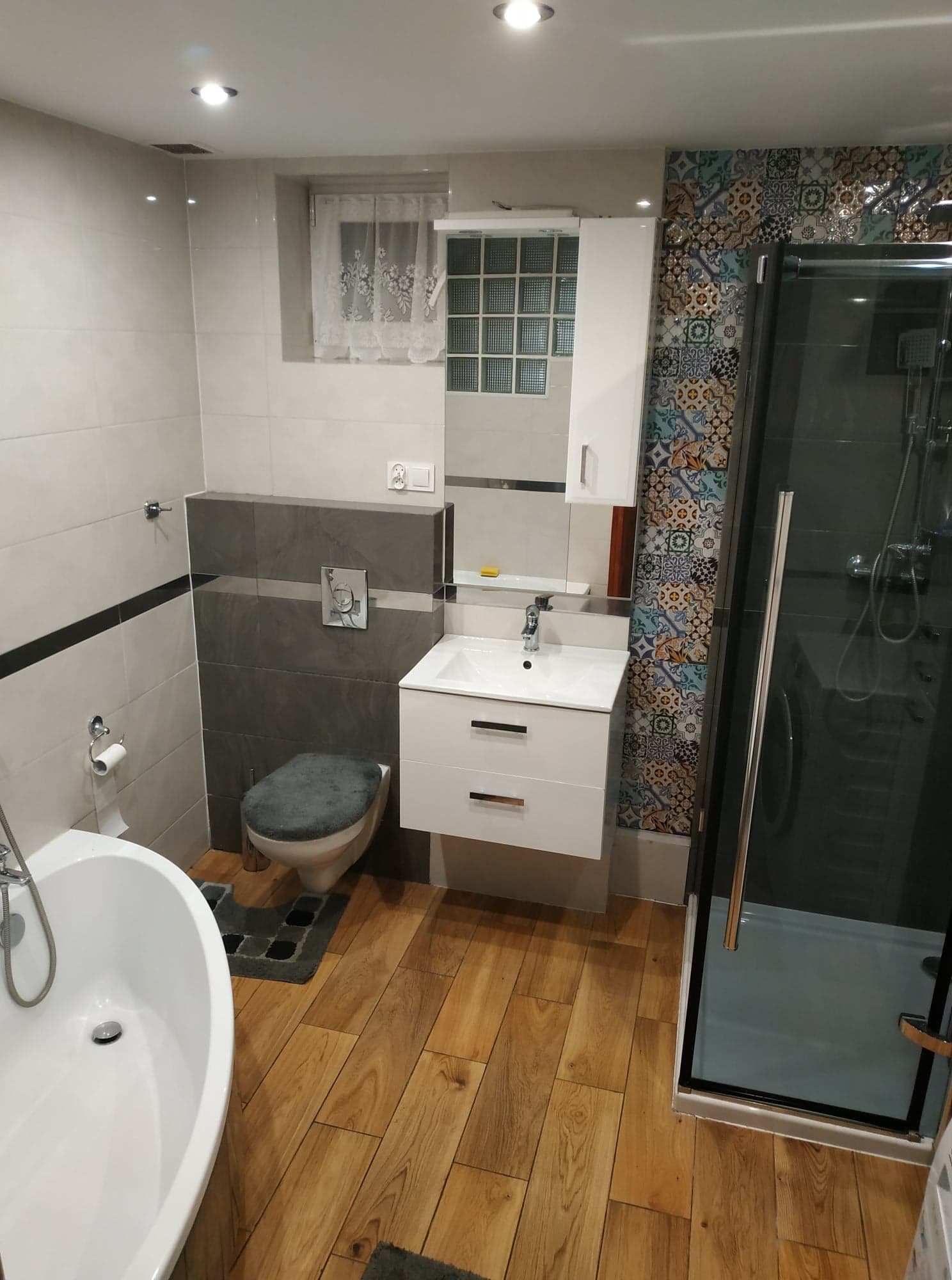 Nowoczesna łazienka po remoncie z drewnopodobną podłogą, białymi płytkami, szarą mozaiką, kabiną prysznicową z czarnymi ramami i białą szafką z umywalką.