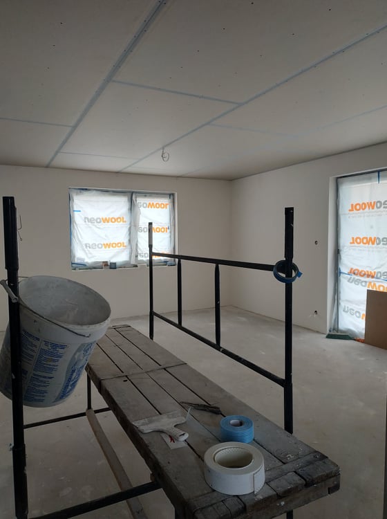 Wnętrze w trakcie prac wykończeniowych: zaklejone okna folią ochronną, sufit z płyt kartonowo-gipsowych z widocznymi łączeniami, rusztowanie z wiadrem i narzędziami.