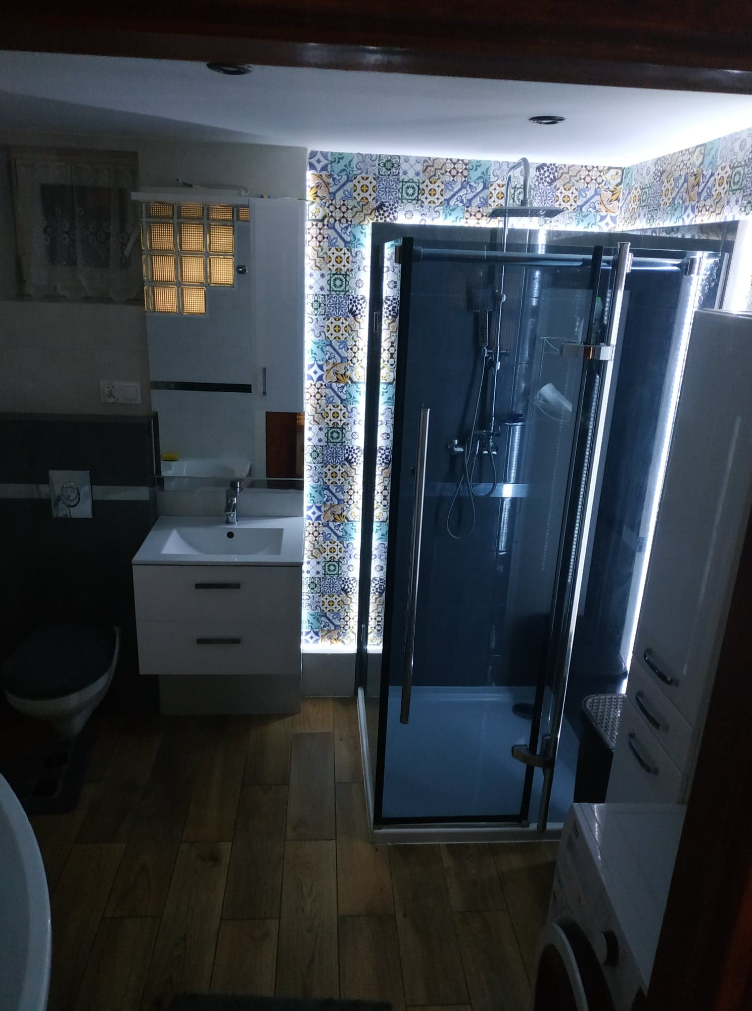 Nowoczesna łazienka z kabiną prysznicową typu walk-in, umywalką z szafką, toaletą i pralką. Ściany wykończone płytkami w geometryczne wzory, podświetlone listwą LED. Drewniana podłoga.