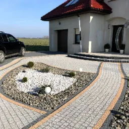 Ułożona kostka granitowa w odcieniach szarości i beżu tworząca ścieżkę do domu z elementami dekoracyjnymi z białych kamieni i niskich krzewów, oświetlona kulistymi lampami, z czarnym SUV-em w tle.