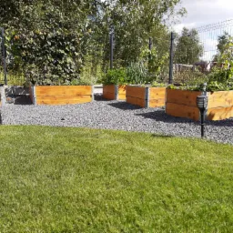 Podwyższone grządki z drewna wypełnione roślinami, otoczone szarym żwirem i trawnikiem, oświetlone lampami solarnymi w ogrodzie z siatkowym ogrodzeniem.