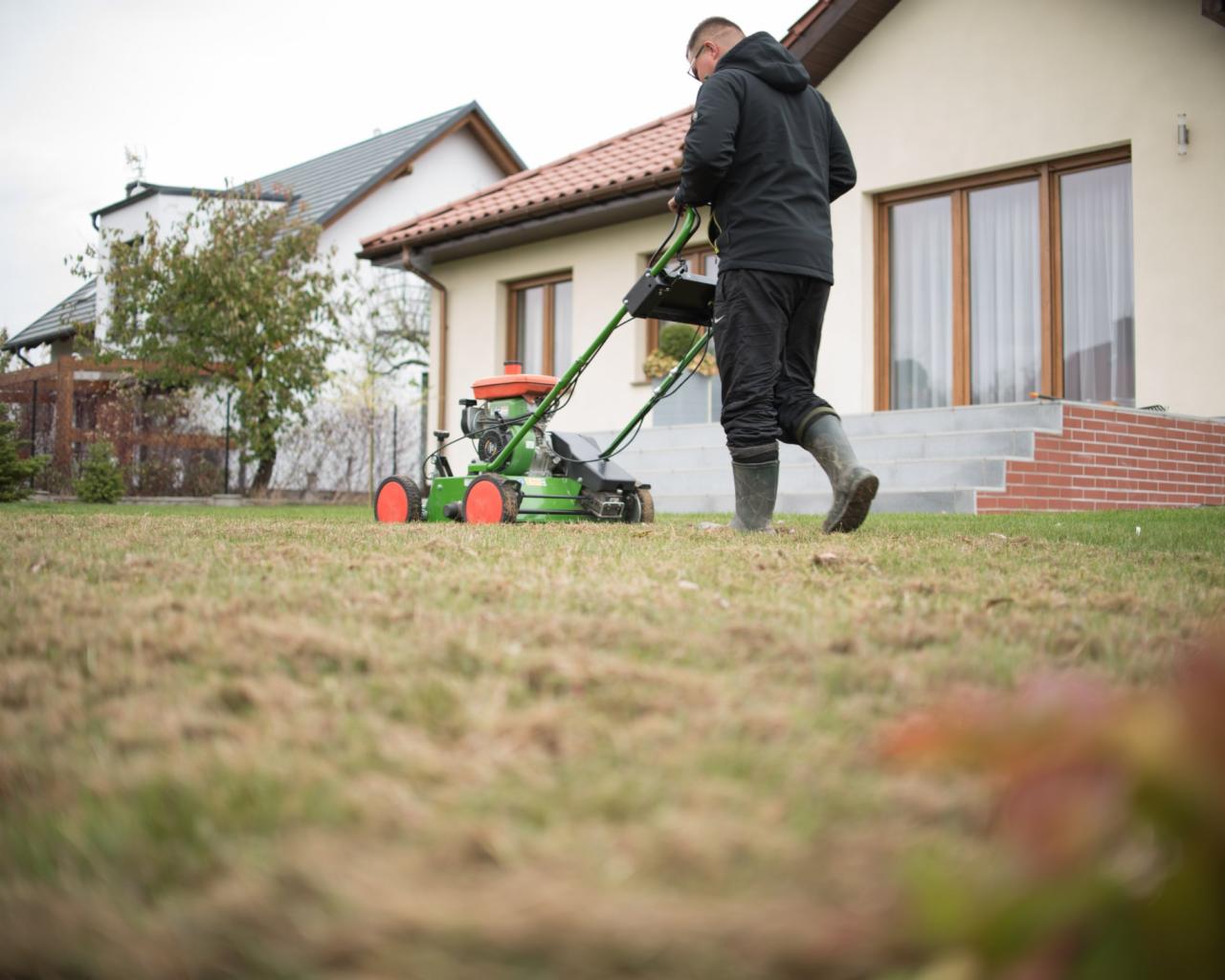 Mężczyzna w gumowcach pcha kosiarkę spalinową po trawniku przed domem jednorodzinnym, widoczne ślady po koszeniu.