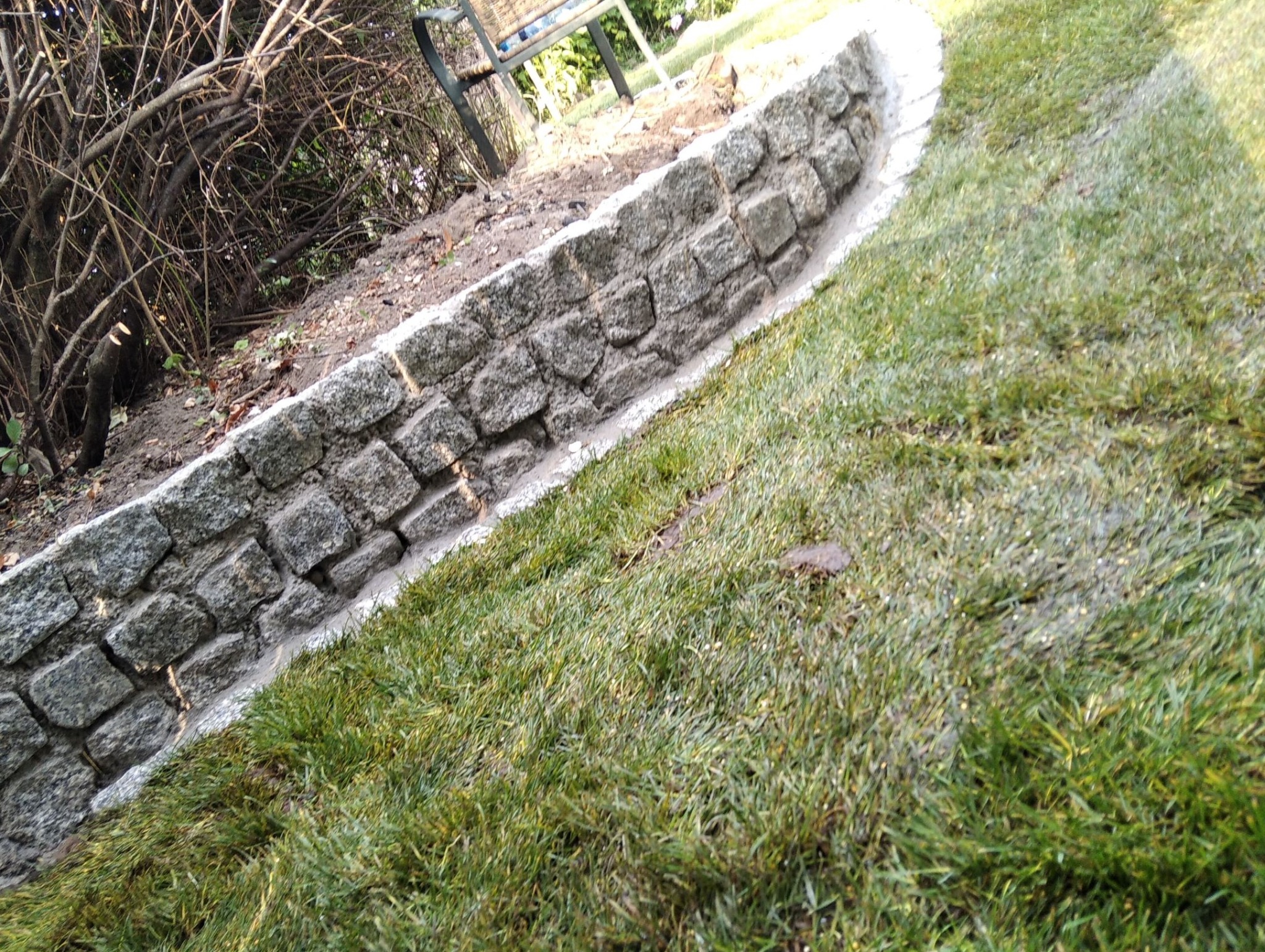 Świeżo ułożona darń przylegająca do niskiego murku z szarego kamienia, widoczne ślady ziemi na trawie.