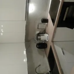 Nowoczesna kuchnia z białymi, połyskującymi frontami szafek, blatem w odcieniu jasnego drewna i marmurowym backsplashiem, wyposażona w czarny zlew, indukcyjną płytę grzewczą, piekarnik oraz ekspres...