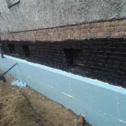 Fundament budynku z nałożoną warstwą hydroizolacji i płytami izolacyjnymi, widoczny wykop i narzędzia.