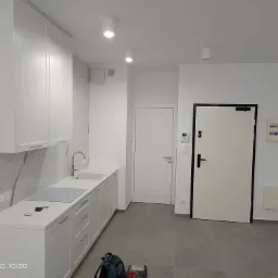 Jasna kuchnia z białymi szafkami, indukcyjną płytą grzewczą i marmurowym backsplashiem; na blacie leżą klucze i karta, a na podłodze torba narzędziowa.