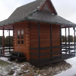 Mały, brązowy domek z drewna z zadaszonym tarasem, kryty ciemnym gontem bitumicznym, stoi na kamiennej podmurówce w zimowym krajobrazie.