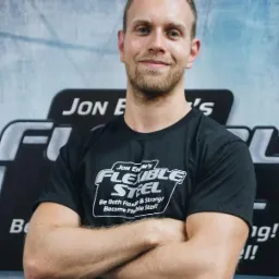 Trener personalny z brodą, w czarnej koszulce z logo 'Flexible Steel', stoi na tle z częściowo widocznym logo tej samej marki, z założonymi rękami i uśmiechem.