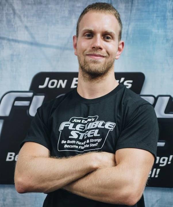 Trener personalny z brodą, w czarnej koszulce z logo 'Flexible Steel', stoi na tle z częściowo widocznym logo tej samej marki, z założonymi rękami i uśmiechem.