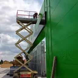 Pracownik na podnośniku koszowym maluje elewację budynku na zielono, widoczna kamera przemysłowa na ścianie.