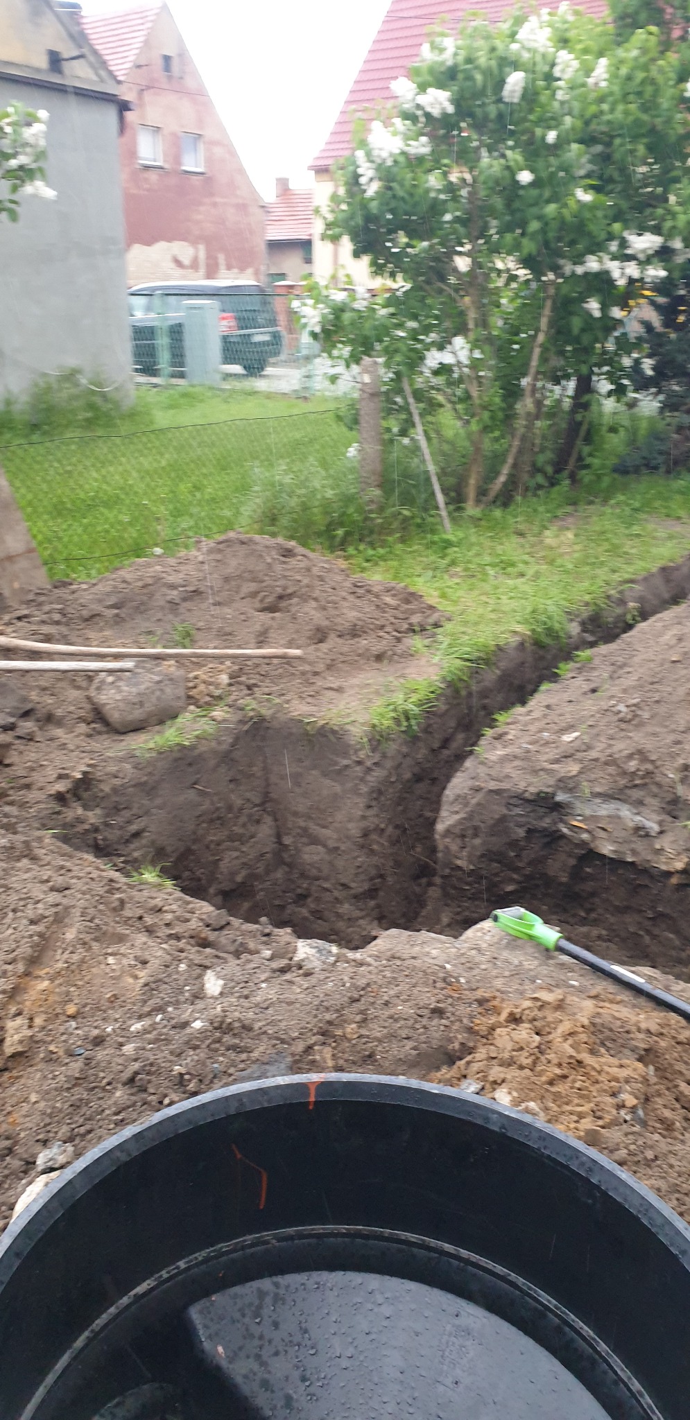 Wykop pod przyłącze wodociągowe do domu jednorodzinnego w trakcie prac ziemnych; widoczny rów, hałda ziemi, fragment studzienki i przewód z zielonym uchwytem.