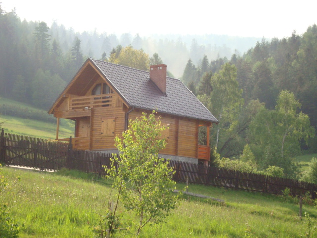 Gotowy, drewniany dom z bali z balkonem, otoczony zielenią i lasem we mgle, widoczny z poziomu trawy za drewnianym ogrodzeniem.