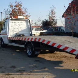 Biała laweta z pomarańczowymi pasami na platformie załadunkowej, zaparkowana na żwirowym podłożu; w tle budynek gospodarczy i drzewa jarzębiny z czerwonymi owocami.