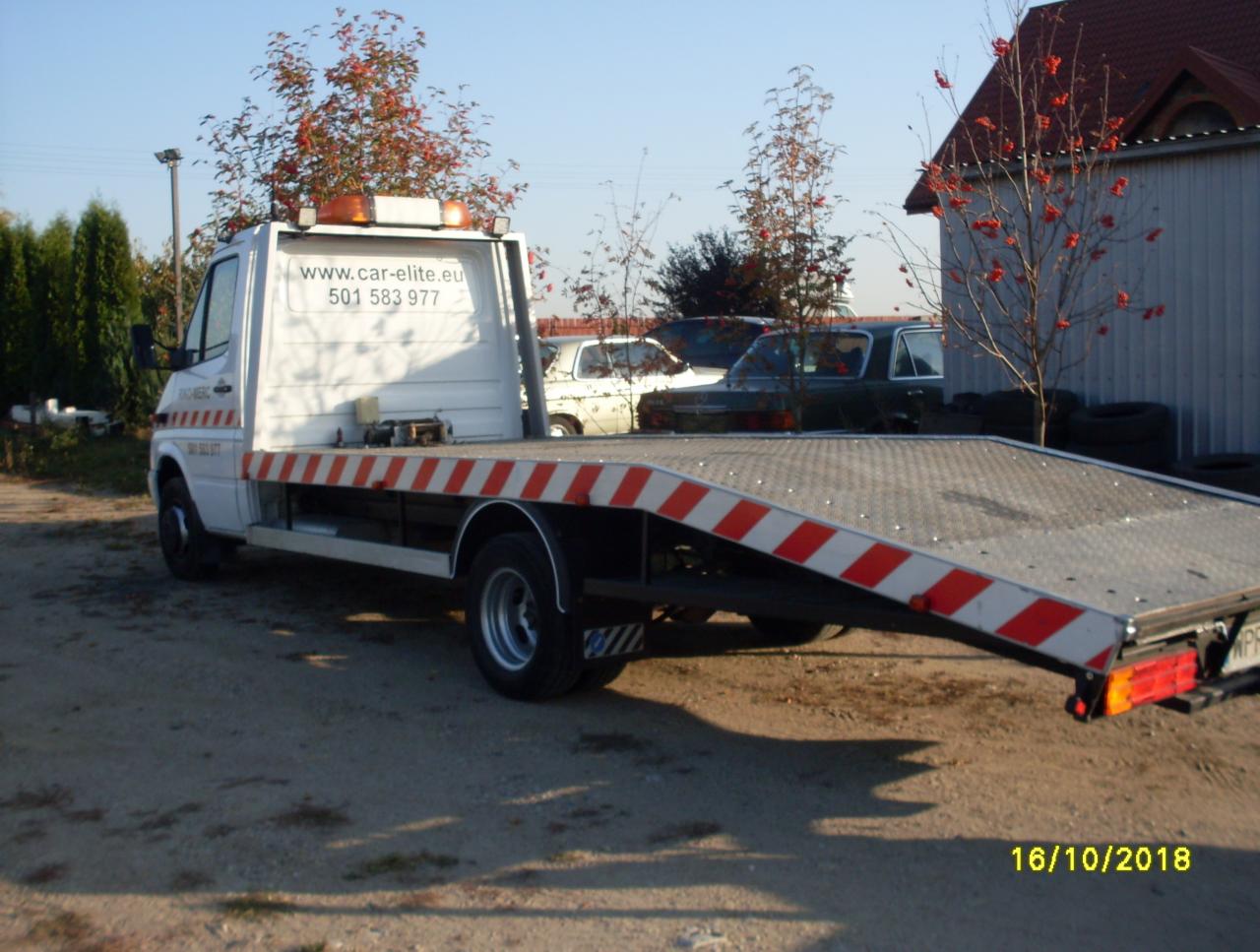 Biała laweta z pomarańczowymi pasami na platformie załadunkowej, zaparkowana na żwirowym podłożu; w tle budynek gospodarczy i drzewa jarzębiny z czerwonymi owocami.