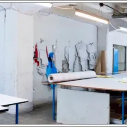 Szeroki kadr hali produkcyjnej z pracownikami układającymi materiał na długich stołach, widoczne rolki materiałów, maszyny do szycia i gotowe elementy odzieży na wieszakach.