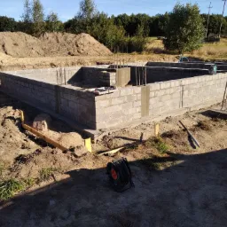 Wzniesione ściany fundamentowe z bloczków betonowych, z wystającymi prętami zbrojeniowymi, na tle pryzmy ziemi i zieleni.