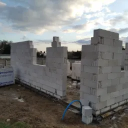 Wznoszenie ścian parteru z białych bloczków betonu komórkowego, widoczne zbrojenie fundamentów, paleta bloczków Prefabet w folii.