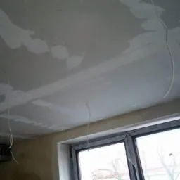 Sufit z płyt kartonowo-gipsowych w trakcie szpachlowania, widoczne przewody elektryczne zwisające z sufitu i fragment okna.