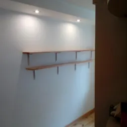 Dwie drewniane półki zamontowane na ścianie z oświetleniem punktowym, widoczne wsporniki mocujące, podłoga z jasnego drewna.