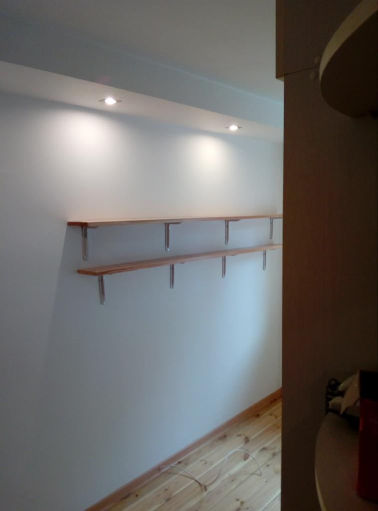 Dwie drewniane półki zamontowane na ścianie z oświetleniem punktowym, widoczne wsporniki mocujące, podłoga z jasnego drewna.