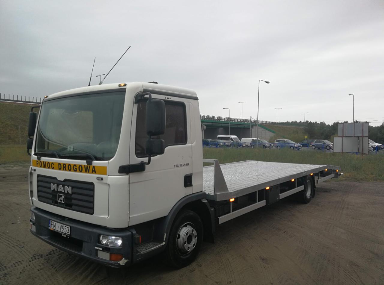 Biała laweta MAN z napisem 'Pomoc Drogowa' na żółtym tle, zaparkowana na piaszczystym terenie, z widocznym fragmentem autostrady w tle.