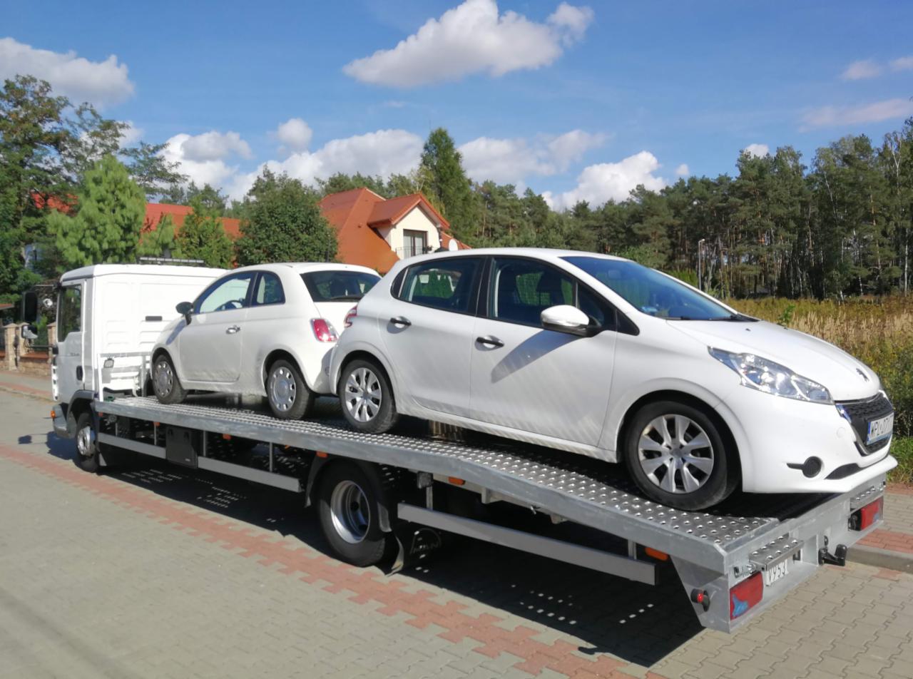 Dwa białe samochody osobowe, Fiat 500 i Peugeot 208, przewożone na platformie lawety w słoneczny dzień.