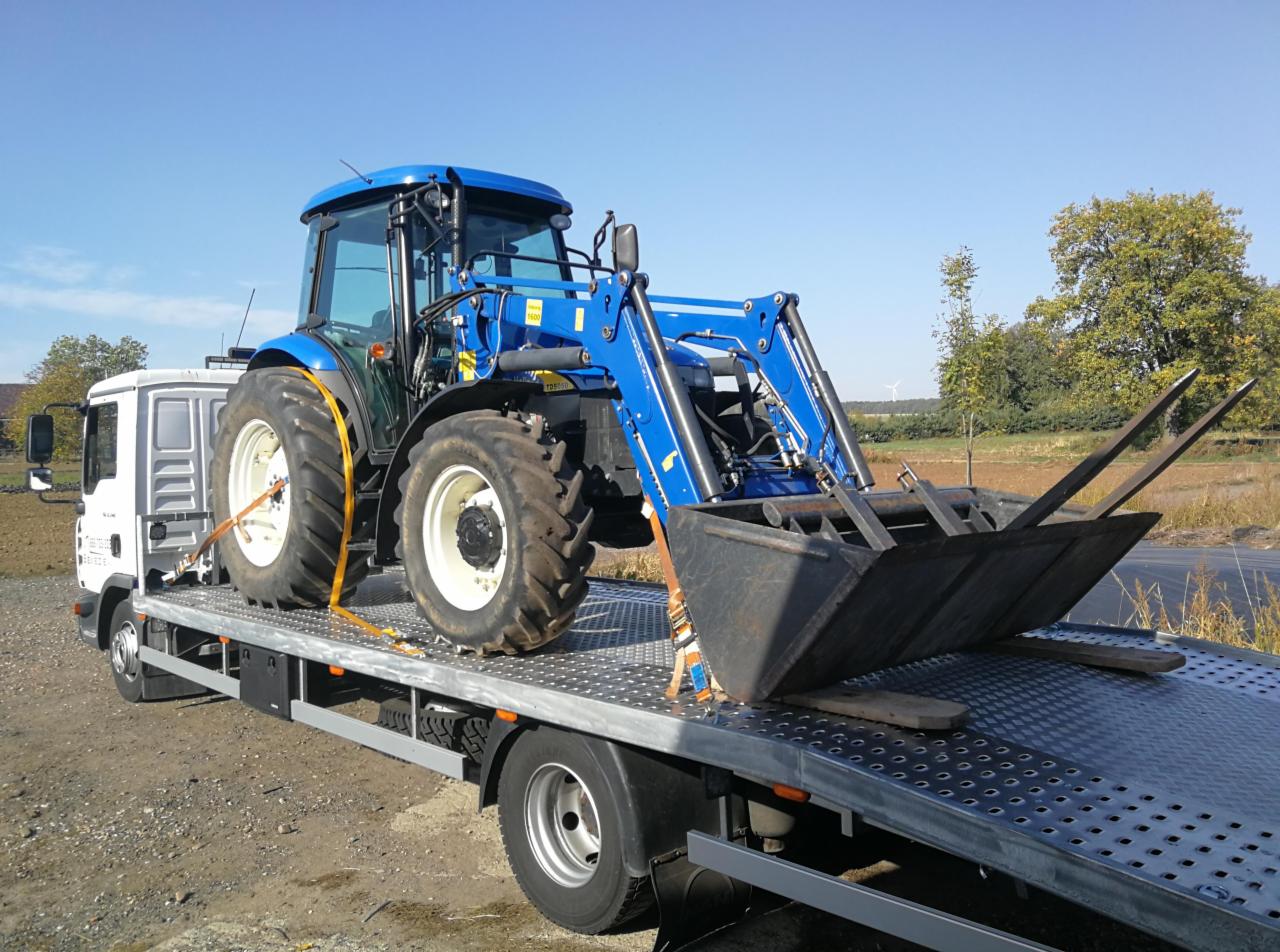 Niebieski traktor z ładowaczem czołowym zabezpieczony pasami transportowymi na platformie lawety, widok z boku, w tle drzewa i błękitne niebo.