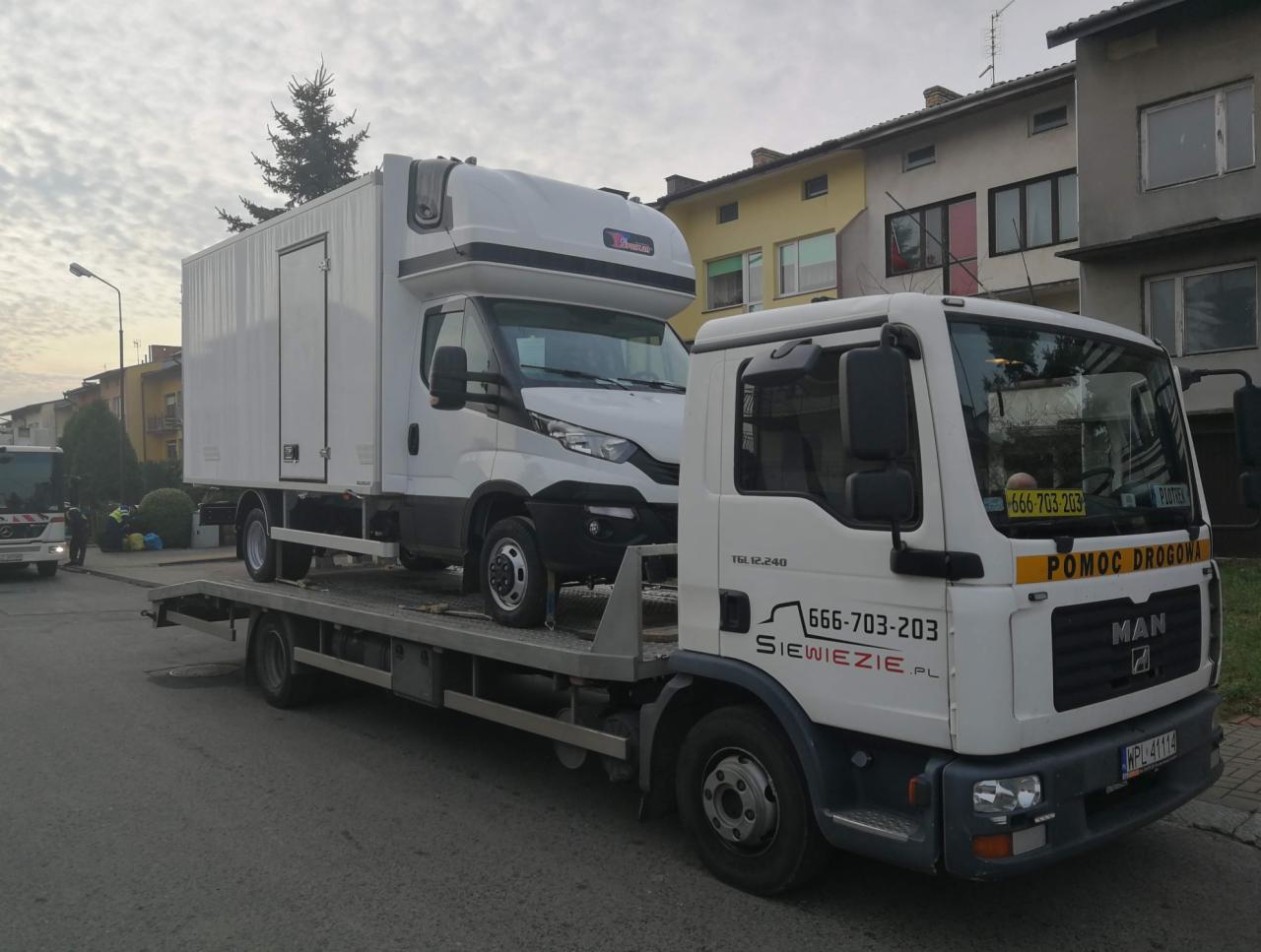 Biała laweta MAN z załadowanym białym busem z zabudową kontenerową na tle zabudowań miejskich.