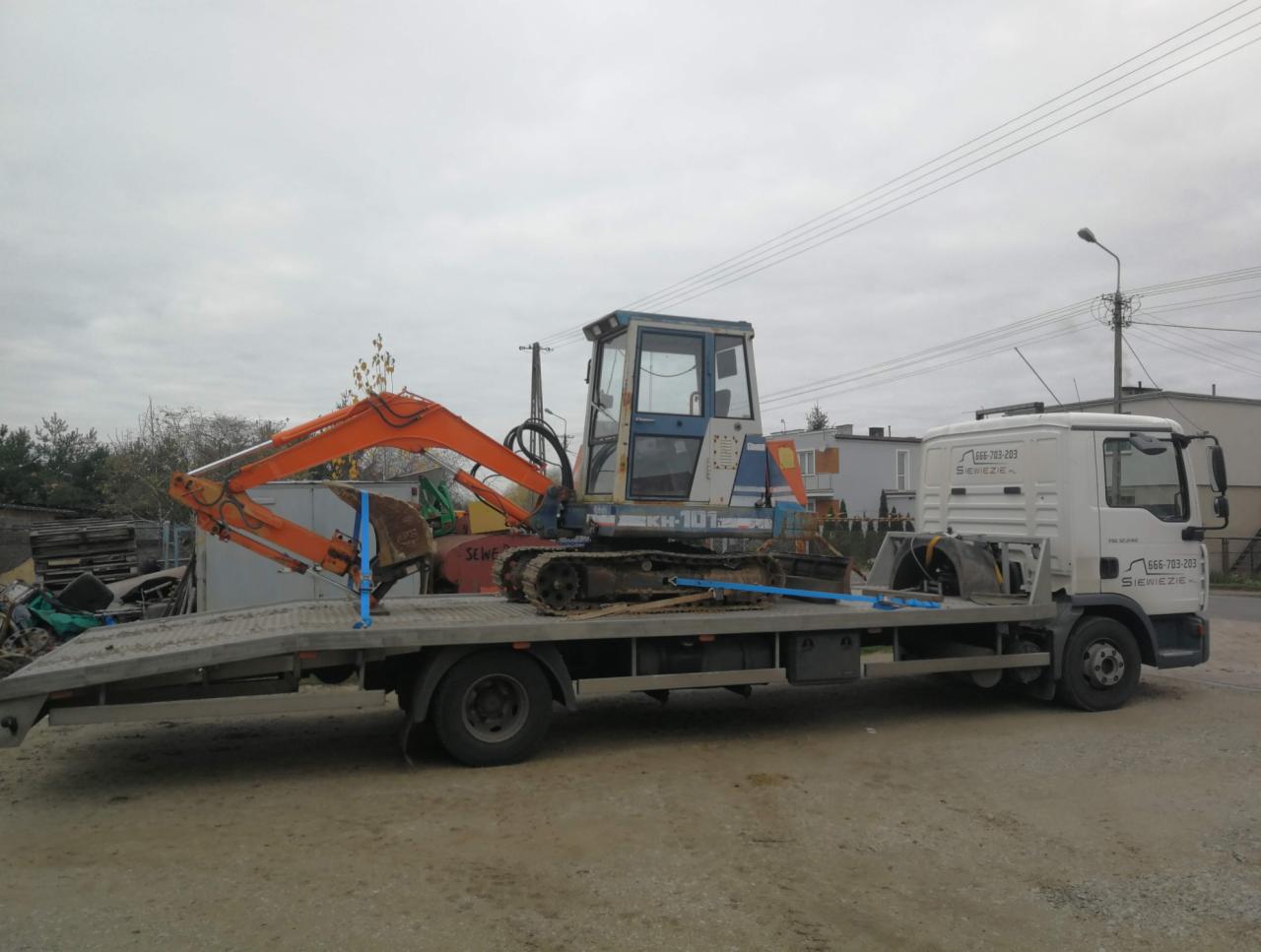 Pomarańczowy minikoparka KH-10 na niskopodwoziowej lawecie z niebieskimi pasami zabezpieczającymi, biała kabina lawety z numerem telefonu i nazwą firmy na tle pochmurnego nieba i zabudowań.