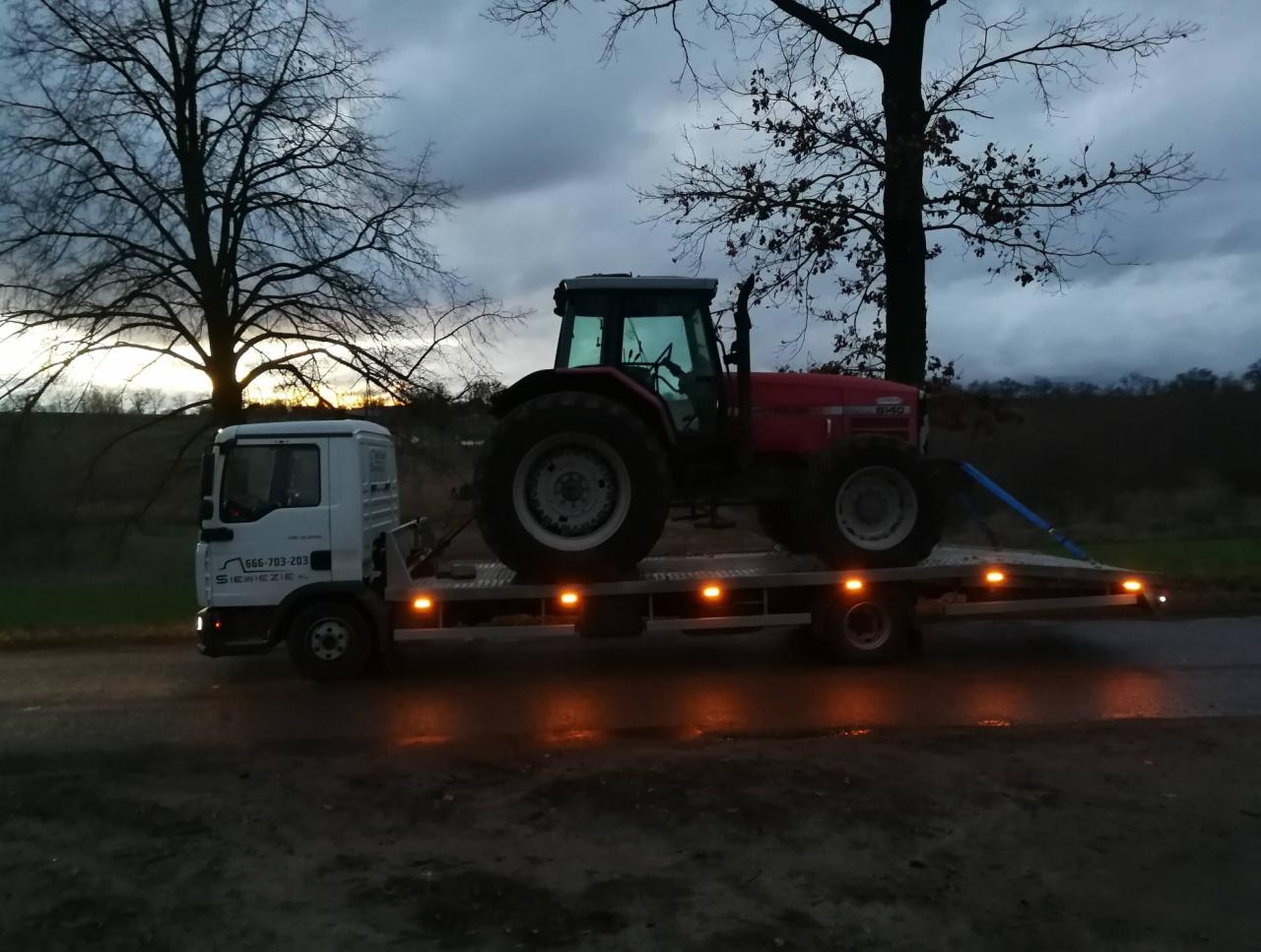 Czerwony traktor zabezpieczony niebieskimi pasami na platformie białej ciężarówki typu laweta, transportowany po asfaltowej drodze o zmierzchu.