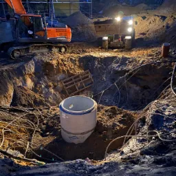 Głęboki wykop oświetlony reflektorami, z widoczną betonową rurą, pomarańczową koparką i traktorem w tle, kable i drewniana paleta w wykopie, budynek w budowie w tle, nocne ujęcie.