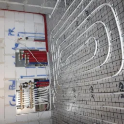 Białe rury ogrzewania podłogowego ułożone na czarnej siatce w trakcie montażu, widoczne także rozdzielacze na ścianie z instalacją wodną.