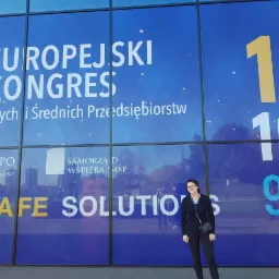 Kobieta pozuje na tle szklanej fasady z napisem 'Europejski Kongres Małych i Średnich Przedsiębiorstw' oraz datami w kolorze niebieskim i żółtym.