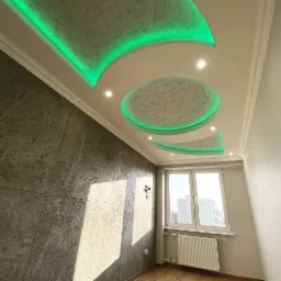 Pokój z dekoracyjnym, podwieszanym sufitem z zielonym oświetleniem LED, ściana z efektem betonu, okno z grzejnikiem.
