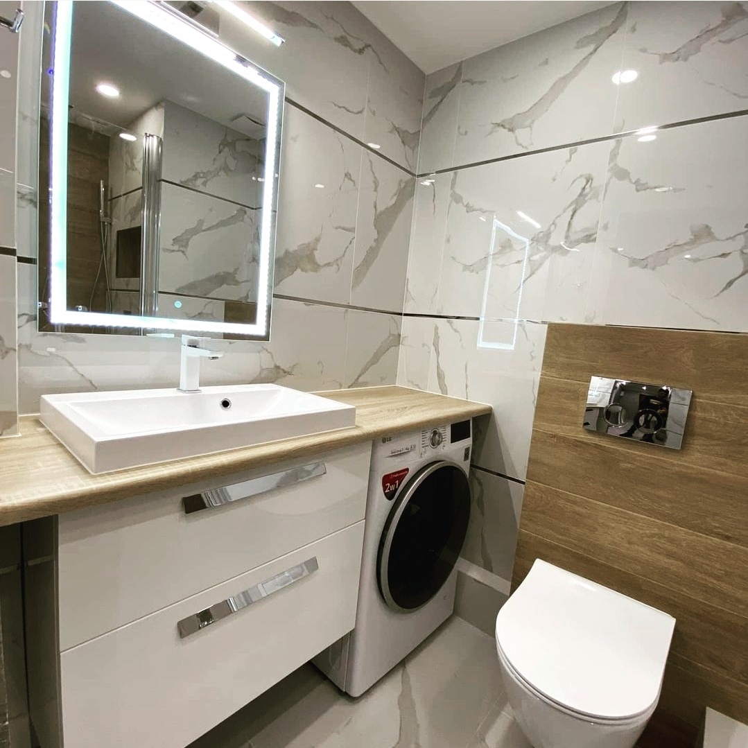Nowoczesna łazienka z marmurowymi płytkami, podświetlanym lustrem, białą umywalką nablatową, szafką z szufladami, pralką LG 2w1 i wiszącą toaletą ze spłuczką w zabudowie z drewnopodobnych paneli.