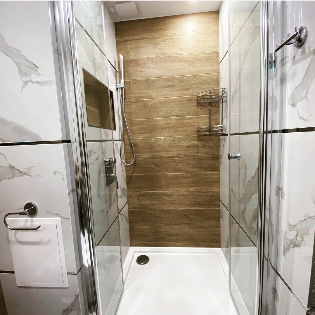 Nowoczesna kabina prysznicowa z białym brodzikiem i szklanymi drzwiami, wykończona płytkami imitującymi marmur i drewno, z chromowaną armaturą i półką na kosmetyki.