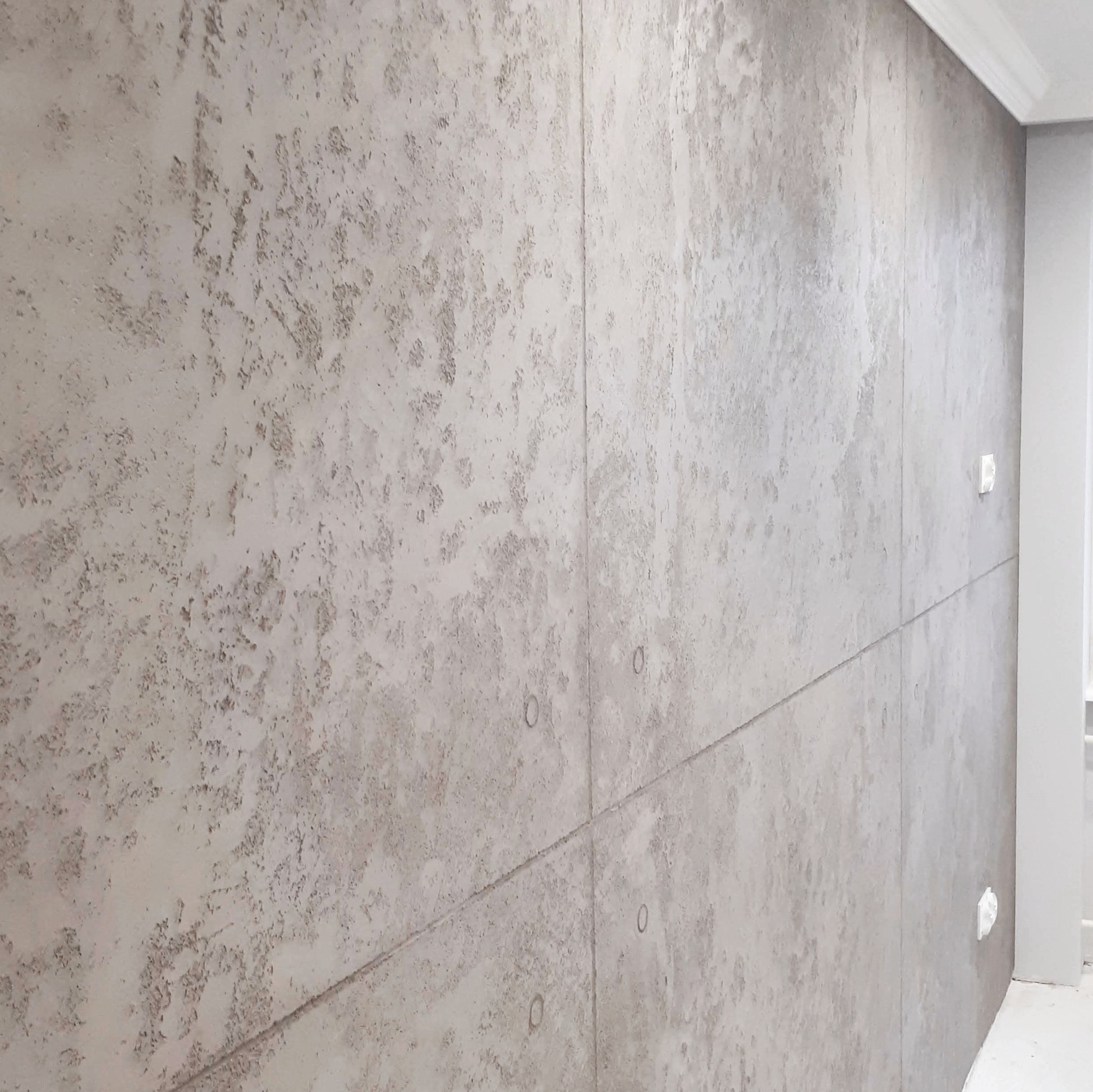 Ściana pokryta szarym tynkiem dekoracyjnym imitującym beton, podzielona na panele z widocznymi spoinami, z zamontowanymi gniazdkami elektrycznymi.