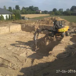 Żółta koparka Mecalac na dnie wykopu pod budowę, widoczne ślady opon i drewniane kołki wyznaczające granice, w tle dom i pole.