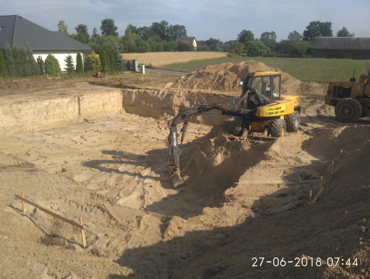 Żółta koparka Mecalac na dnie wykopu pod budowę, widoczne ślady opon i drewniane kołki wyznaczające granice, w tle dom i pole.