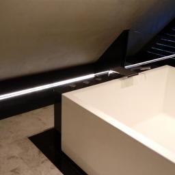 ALAN - Nowoczesna biała wanna prostokątna z chromowaną armaturą, podświetlenie LED wzdłuż ściany, czarno-beżowa marmurowa posadzka w łazience na poddaszu.