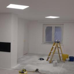 ALAN - Pokój w trakcie remontu: świeżo pomalowane na biało ściany, zabezpieczone okno folią, drabina malarska, wiadro z farbą i akcesoria malarskie na podłodze pokrytej folią ochronną, czarny wkład...