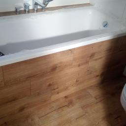 ALAN - Biała wanna zabezpieczona folią ochronną, z zamontowaną obudową z płytek imitujących drewno, widoczny fragment podłogi z tego samego materiału oraz toaleta w trakcie remontu łazienki.