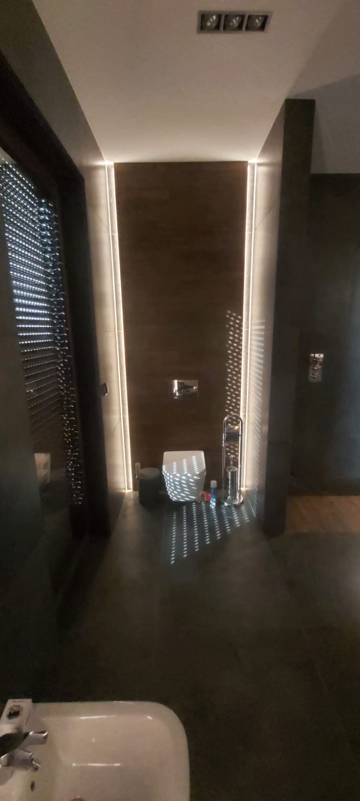 Nowoczesna łazienka z brązowymi płytkami, podświetleniem LED wzdłuż ściany i ciemną podłogą. Na podłodze widoczne odbicia światła tworzące wzór. Umywalka z bidetem w dolnej części kadru.