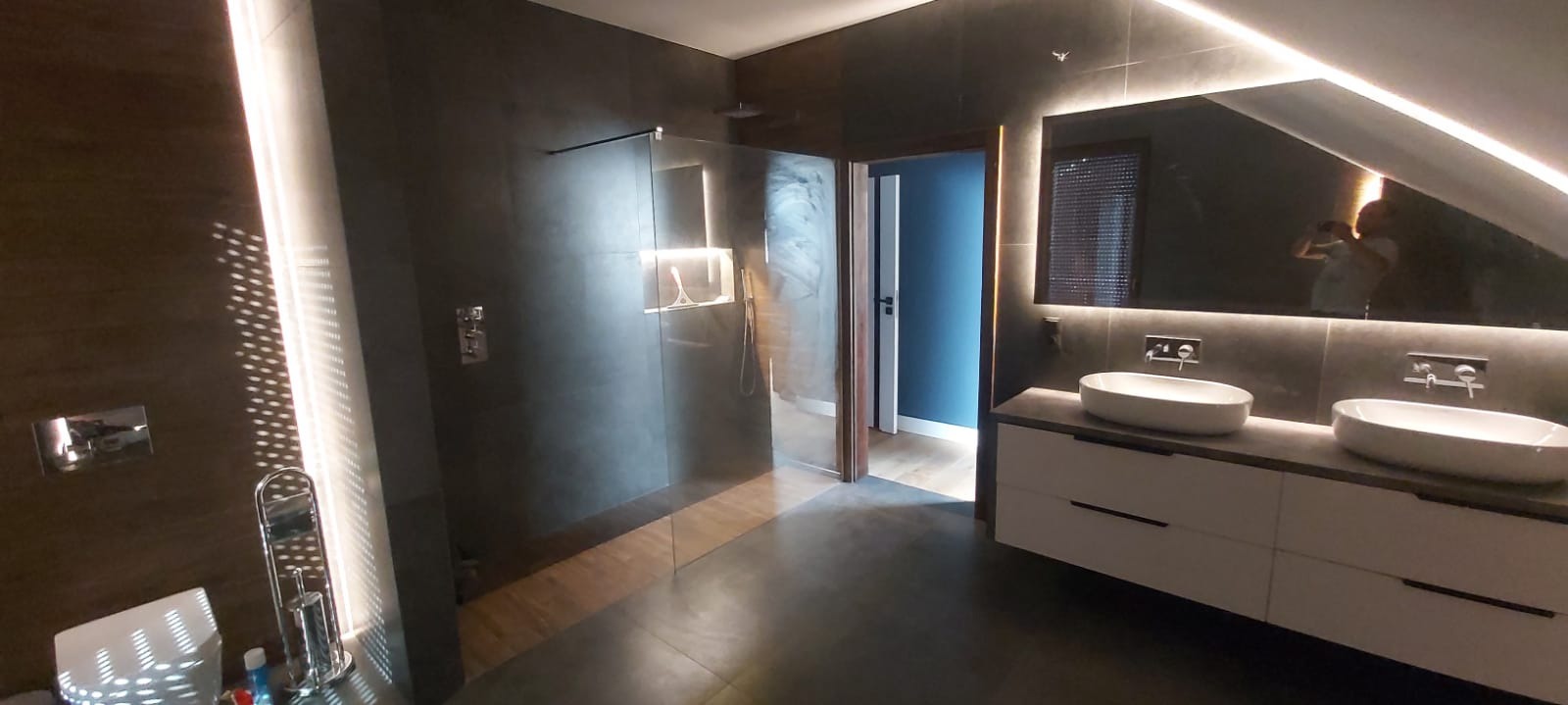 Nowoczesna łazienka z ciemnymi płytkami, kabiną prysznicową typu walk-in i podwójną umywalką nablatową, oświetleniem LED i widocznym odbiciem osoby robiącej zdjęcie w lustrze.