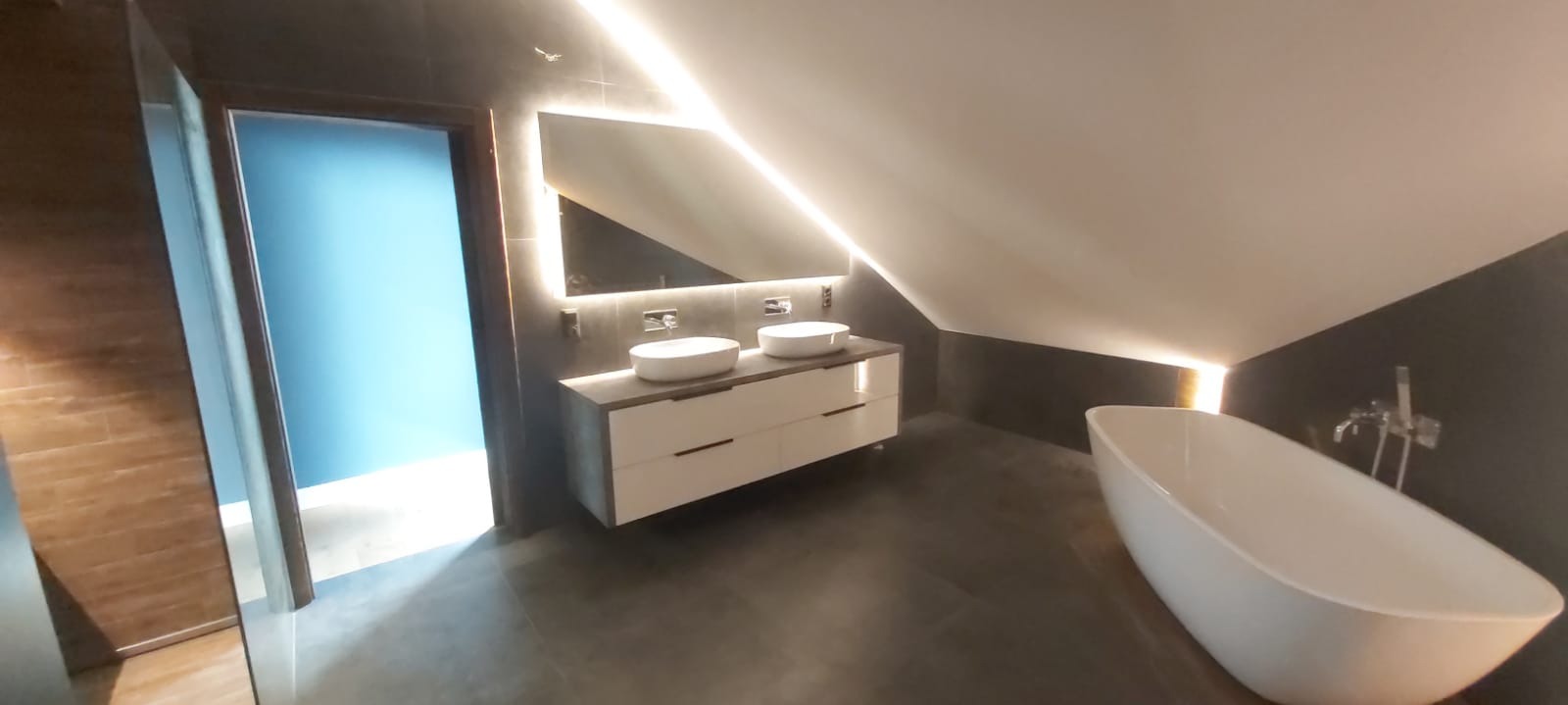 Nowoczesna łazienka na poddaszu z dwoma umywalkami nablatowymi, podświetlanym lustrem, wolnostojącą wanną i drzwiami do innego pomieszczenia z niebieską ścianą w tle.