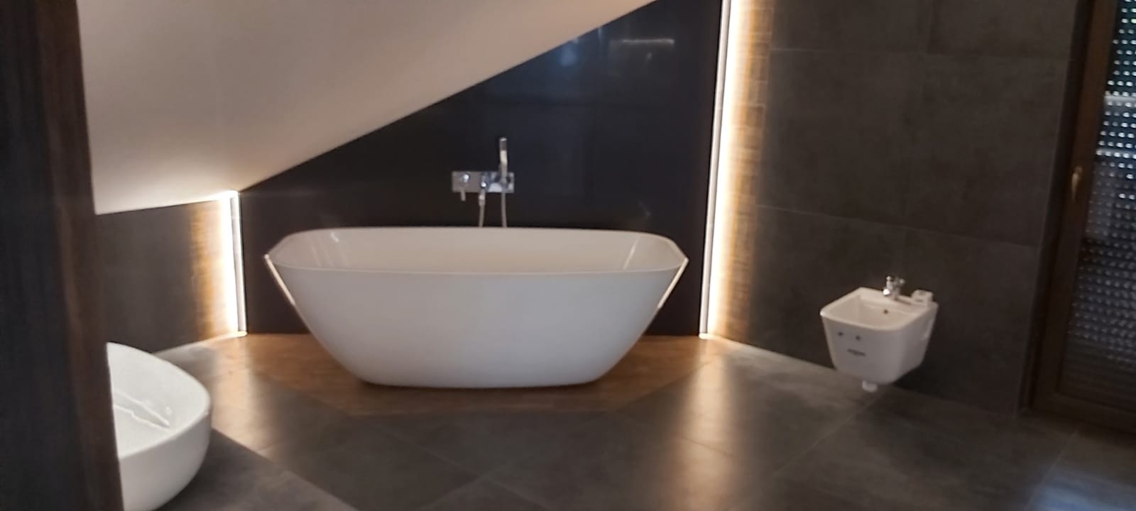 Nowoczesna łazienka na poddaszu z wolnostojącą wanną, bidetem i szarą glazurą, oświetlona listwami LED.