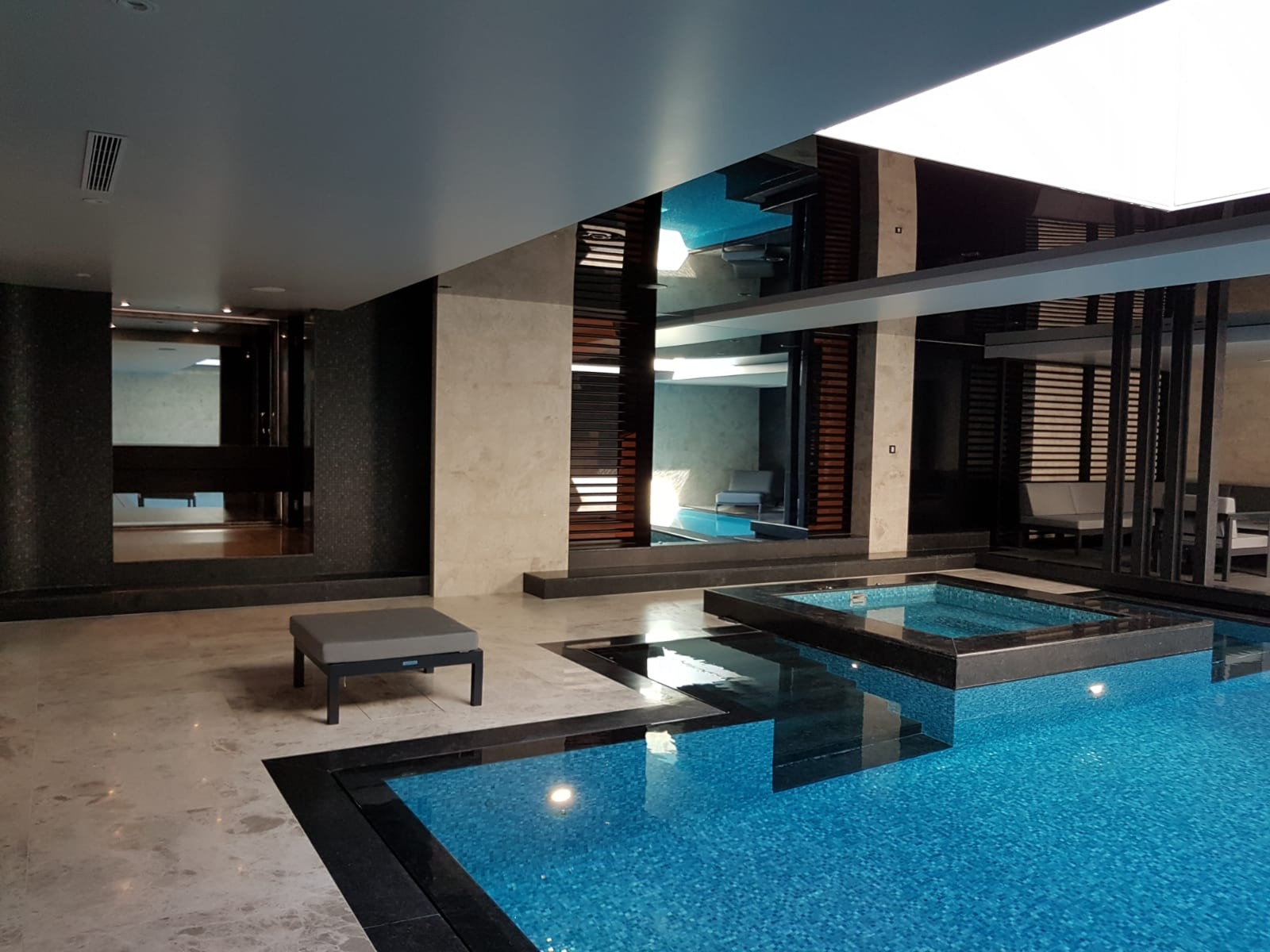Wnętrze nowoczesnego basenu z niebieską mozaiką, szarym siedziskiem i lustrzanymi elementami na ścianach.